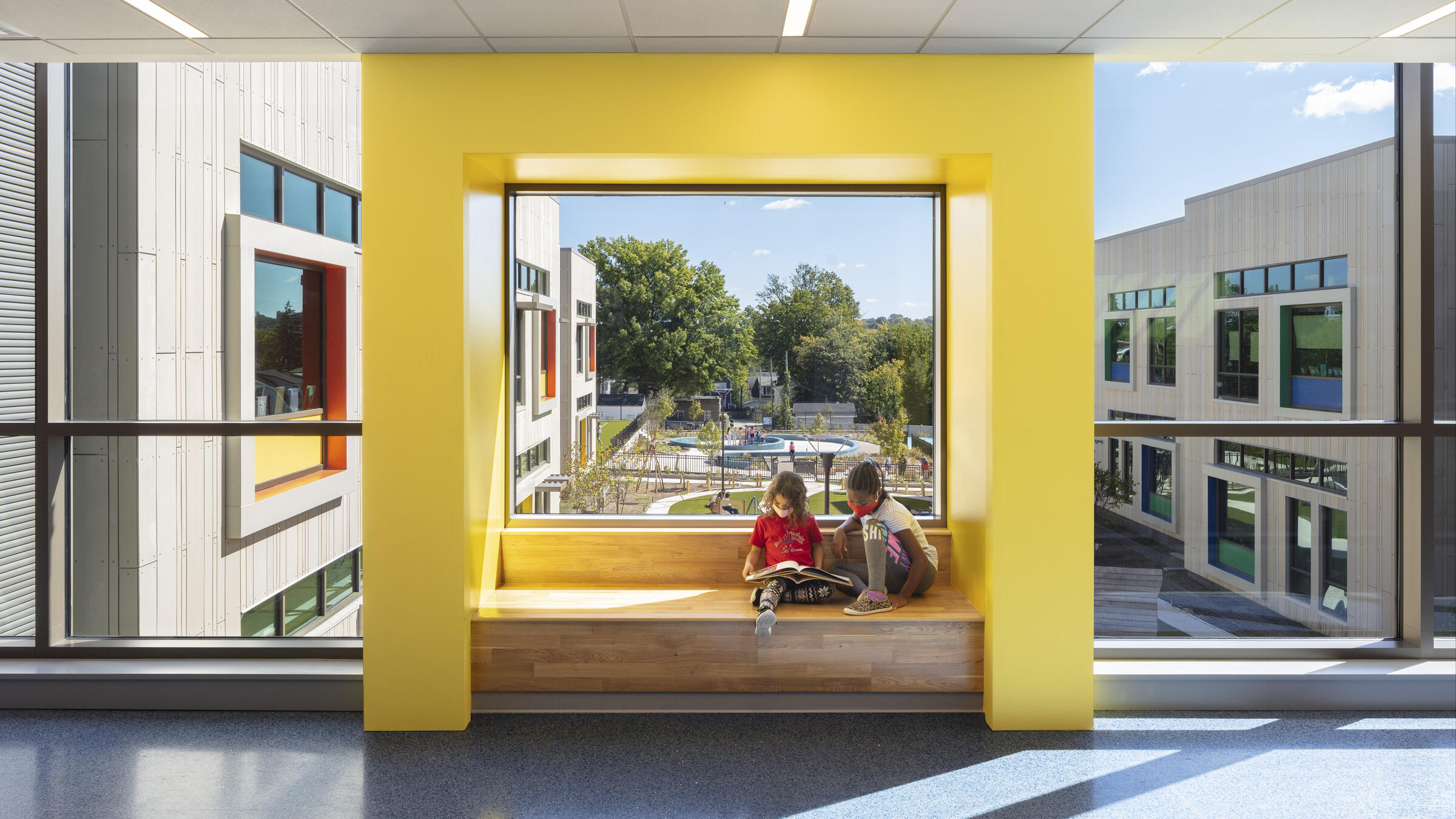 Children read a book on a bench in a brightly lit, sunny hallway of John Lewis Elementary School.